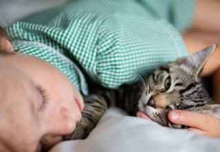 A child in a green shirt lying on a bed while holding a tabby cat.