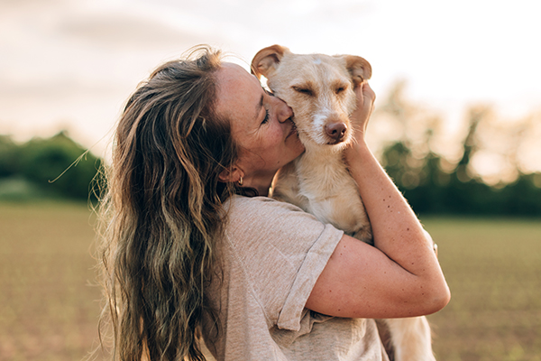 A woman holds and kisses a small, scruffy tan dog.